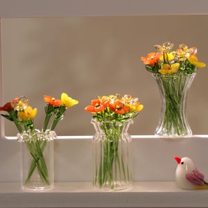 May include: Glass flower sculptures in clear vases. The flowers are orange, yellow, and clear, with green stems. The vases have different shapes and sizes. A small, white bird figurine with pink and yellow accents is also present.
