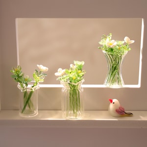 Pode incluir: Três vasos de vidro com arranjos de flores em miniatura e uma pequena estatueta de pássaro. Os vasos são transparentes e contêm delicadas flores verdes e brancas. O pássaro é branco com detalhes em vermelho e rosa.