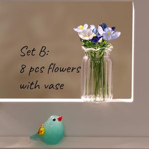 May include: A clear glass vase with a ruffled top holds eight small flowers in shades of white, blue, and yellow. The vase sits on a white surface, and a small, turquoise glass bird with a red beak is in the foreground. Text reads "Set B: 8 pcs flowers with vase."