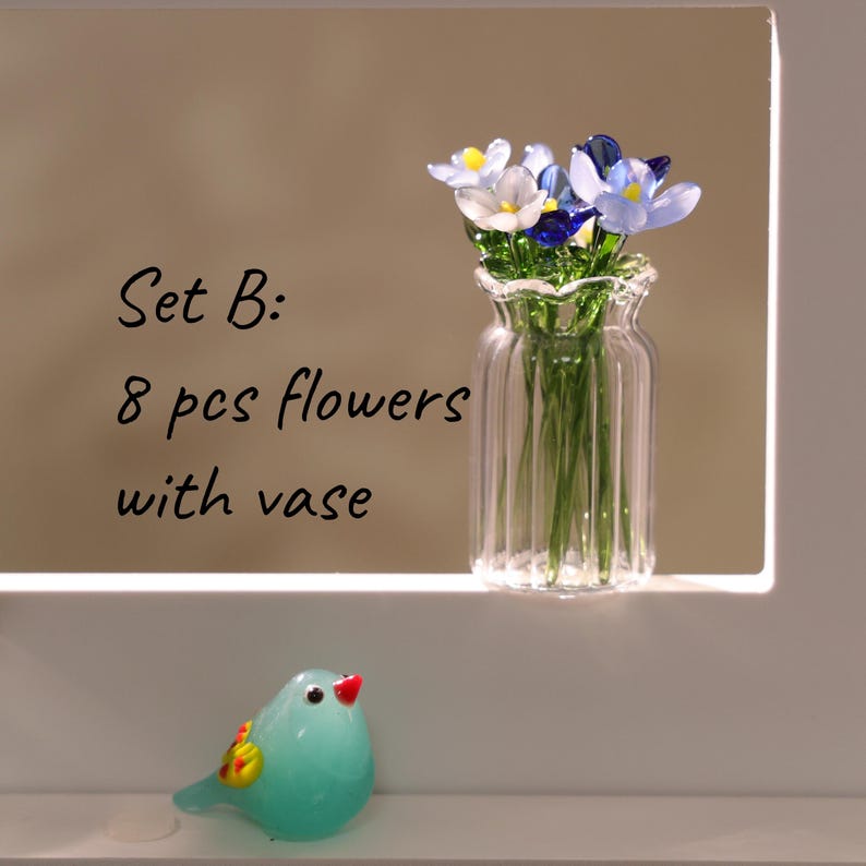 May include: A clear glass vase with a ruffled top holds eight small flowers in shades of white, blue, and yellow. The vase sits on a white surface, and a small, turquoise glass bird with a red beak is in the foreground. Text reads "Set B: 8 pcs flowers with vase."