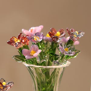 May include: A glass flower bouquet in a clear vase. The flowers are various colors, including pink, purple, and red, with yellow centers. The stems and leaves are green. The vase has a flared rim.