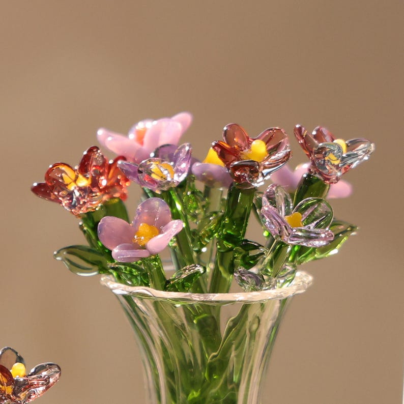 May include: A glass flower bouquet in a clear vase. The flowers are various colours, including pink, purple, and red, with yellow centres. The stems and leaves are green. The vase has a flared rim.