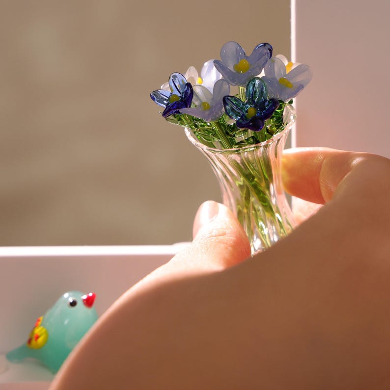 May include: A miniature glass vase filled with delicate glass flowers in shades of blue and white. The vase is held by a person's hand. A small, turquoise glass bird with red and yellow accents is in the background.