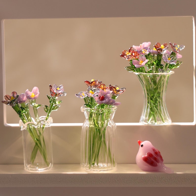 May include: Glass flower bouquets in clear vases with pink and orange flower accents. A small, pink glass bird figurine is also present. The vases have ruffled edges and ribbed detailing.