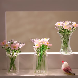 May include: Three glass vases with pink and yellow flower arrangements. The vases are clear and have different designs. A small, decorative bird figurine with red, white, and pink details sits nearby.