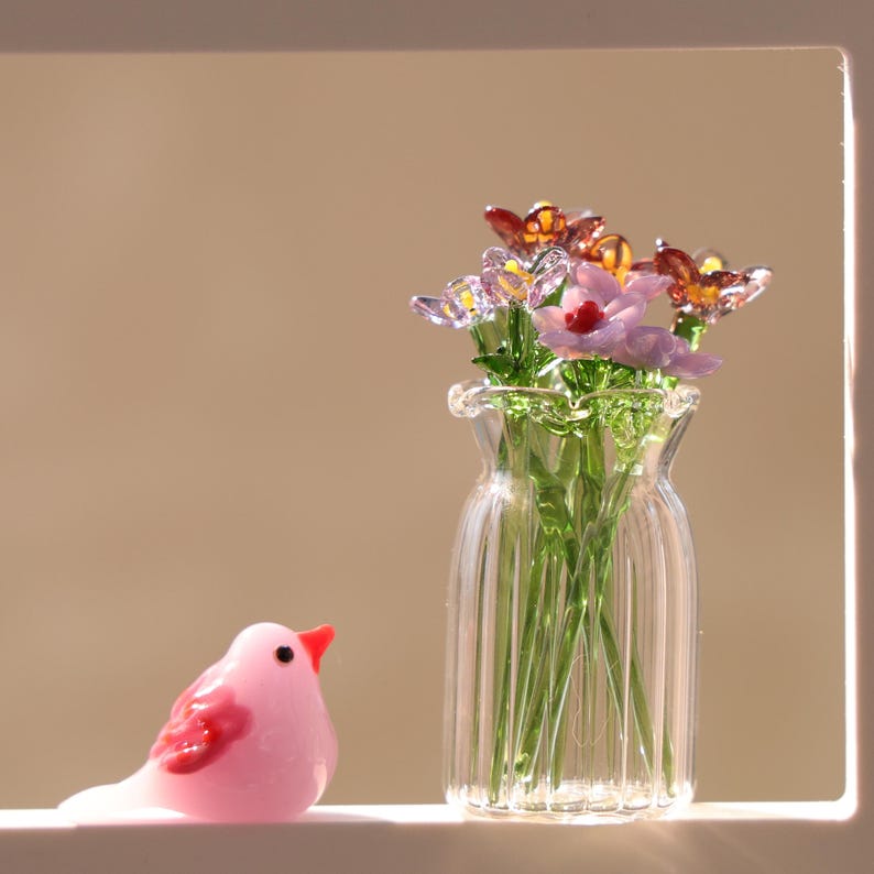 May include: A clear glass vase with a ruffled rim contains delicate glass flowers in shades of purple, pink, and orange. A small, pink bird figurine with red accents sits to the left of the vase, creating a charming decorative scene.