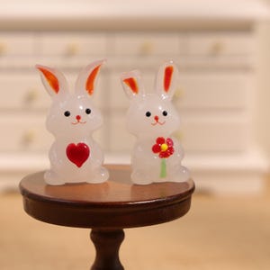 Puede incluir: Dos figuras translúcidas de conejos blancos con orejas con puntas naranjas y detalles rojos. Uno tiene un corazón rojo, el otro sostiene una flor roja y amarilla. Están sobre una pequeña mesa redonda de madera.