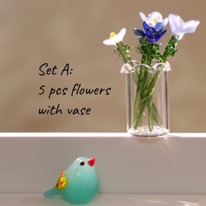 May include: A glass vase holding five colorful glass flowers with green stems. The flowers are white, blue, and purple. A small, turquoise glass bird with a red beak and yellow and orange accents sits in front of the vase. Text on the left reads "Set A: 5 pcs flowers with vase."