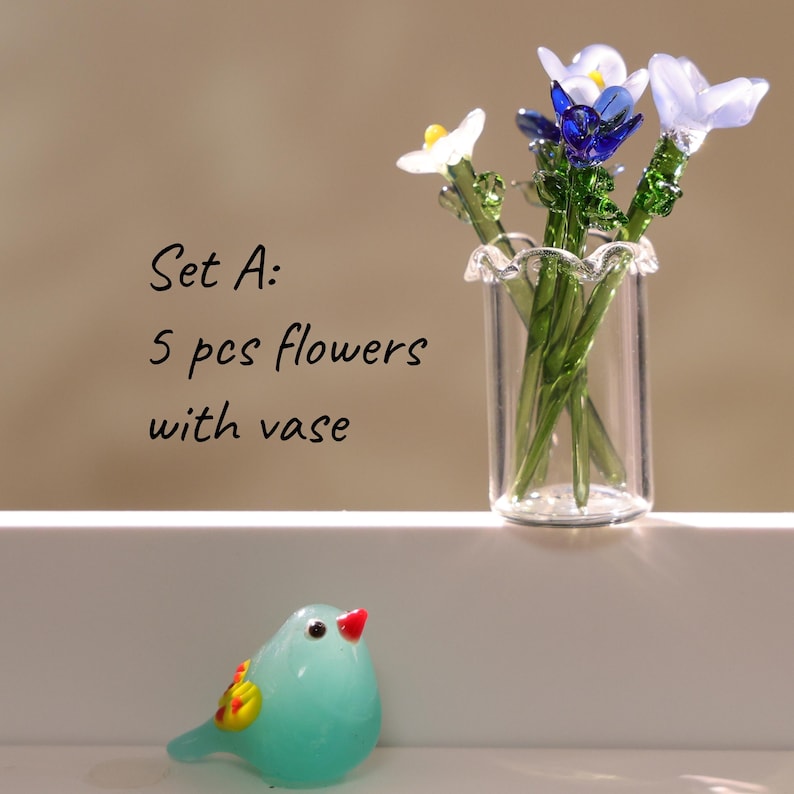 May include: A glass vase holding five colorful glass flowers with green stems. The flowers are white, blue, and purple. A small, turquoise glass bird with a red beak and yellow and orange accents sits in front of the vase. Text on the left reads "Set A: 5 pcs flowers with vase."