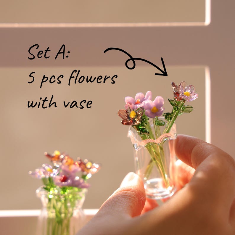 May include: A small glass vase containing a miniature bouquet of flowers in shades of pink, purple, and yellow, with green stems. The text "Set A: 5 pcs flowers with vase" is visible.