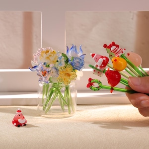 Può includere: Un vaso di vetro trasparente pieno di fiori di vetro colorati, tra cui fiori gialli, blu e bianchi. Una piccola statuina rossa di Babbo Natale è vicina. Una mano tiene diversi stuzzicadenti decorativi in vetro con disegni festivi.