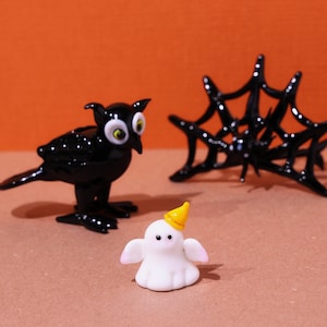 May include: A Halloween-themed display featuring a black crow figurine with large yellow eyes, a white ghost with a yellow party hat, and a black spider web. The figurines are set against an orange background.
