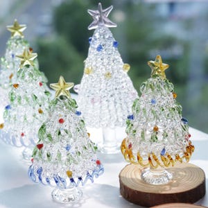 May include: A collection of decorative glass Christmas trees. Each tree is clear with colorful accents, including green, blue, red, and yellow. The trees are topped with star ornaments and sit on clear bases or a wooden slice.