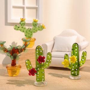 以下が含まれることがあります： ミニチュアガラス製のサボテン彫刻のコレクション。サボテンはさまざまな緑色で、白い点と赤または黄色の花のアクセントが付いています。小さな黄色の鉢に飾られています。