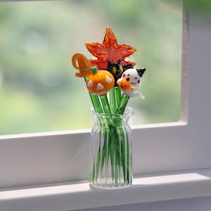 Puede incluir: Un jarrón de vidrio transparente contiene una colección de figuras de vidrio con temática de Halloween. Las figuras incluyen una calabaza, un gato negro, un fantasma, una hoja de arce y un sombrero de bruja. Las figuras están unidas a tallos verdes.