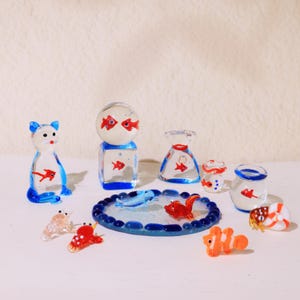 Peut inclure: Collection de figurines miniatures en verre, dont un chat bleu et blanc, un cube surmonté d'une sphère et un étang avec des poissons. Les figurines sont transparentes avec des accents rouges et bleus, représentant des poissons et la vie aquatique.