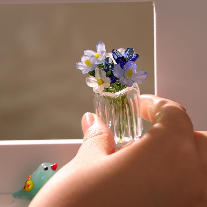 May include: A miniature glass vase filled with delicate, colorful glass flowers. The flowers feature white, light blue, and dark blue petals with yellow centers. The vase is held in a hand, showcasing its small size and intricate details. A small glass bird is in the background.