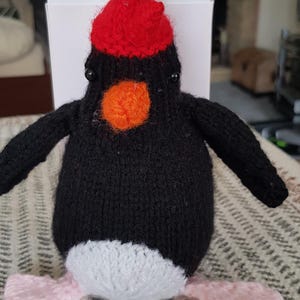 以下が含まれることがあります： 赤いニット帽をかぶった黒と白のニットのペンギン。ペンギンは明るいオレンジ色のくちばしと黒い目をしています。ペンギンは模様のある敷物の上に座っています。
