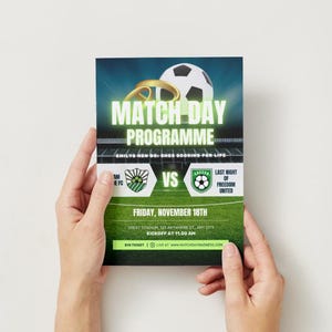 May include: A match day programme held by hands, featuring a soccer ball and gold rings. The green and white design includes team logos, the date, and the time of the match. The text reads "MATCH DAY PROGRAMME".