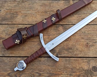 May include: A medieval sword with a silver blade and a brown leather-wrapped handle, lying next to its brown leather sheath. The sword's crossguard has the word "CHRISTUS" and "VINCIT" etched on it, and the sheath is adorned with gold crosses.