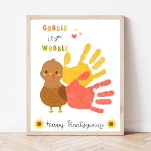 May include: Thanksgiving-themed art print featuring a cartoon turkey, handprints, and the phrase "Gobble til you Wobble." The print includes sunflowers and the text "Happy Thanksgiving" in a wooden frame.