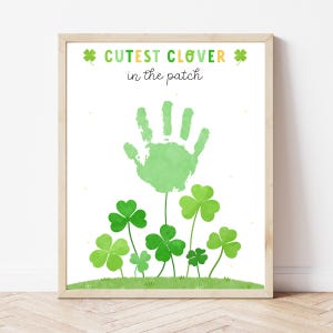 May include: A framed print with a light wood frame. The print features a green handprint above a field of green clovers. The text "CUTEST CLOVER in the patch" is written in green and yellow.
