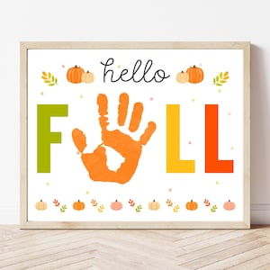 Op de afbeelding: Een ingelijste print met de woorden "hello FALL" in een speels lettertype. De "F" is groen, de handafdruk is oranje en de "LL" is geel en oranje. Pompoenen en bladeren versieren de boven- en onderkant.
