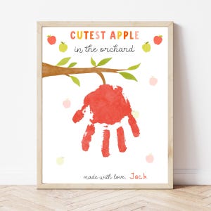May include: A framed print with the text "Cutest Apple in the orchard." The artwork features a red handprint hanging from a tree branch, with small apple illustrations. The print is signed "made with love, Jack."