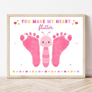 Peut inclure: Une impression encadrée avec un motif de papillon rose fait avec des empreintes de pieds. Le papillon a un corps rose et des ailes faites d'empreintes de pieds. Le texte "YOU MAKE MY HEART flutter" est au-dessus du papillon, avec des petits coeurs.