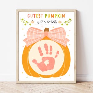 May include: A framed print featuring a watercolor pumpkin design. The pumpkin has a handprint in the center and a pink bow on top. The text reads "Cutest Pumpkin in the Patch" in colorful letters.