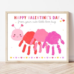 Op de afbeelding: Een ingelijste print met de tekst "HAPPY VALENTINE'S DAY" en "from your cute little love bug". Het kunstwerk toont een roze handafdrukrups met een lachend gezicht en hartantennes. De print heeft een witte achtergrond met een rand van harten.