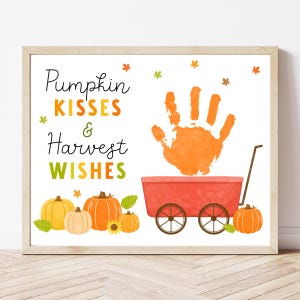 Könnte beinhalten: Gerahmter Druck mit dem Text "Pumpkin Kisses & Harvest Wishes". Das Kunstwerk zeigt einen orangefarbenen Handabdruck, einen roten Wagen und mehrere Kürbisse in Orange- und Gelbtönen. Dekorative Herbstblätter sind verstreut.