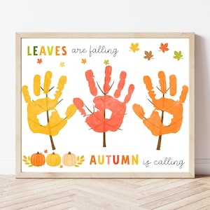 Puede incluir: Una impresión enmarcada con las palabras "LEAVES are falling" y "AUTUMN is calling". La obra de arte presenta tres huellas de manos en forma de hojas en tonos amarillos, naranjas y rojos, con pequeñas calabazas y detalles de hojas.