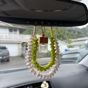 May include: Car air freshener with a white and green floral design, hanging from the rearview mirror. The freshener has a small glass bottle and a wooden top. A white flower is clipped to the dashboard.