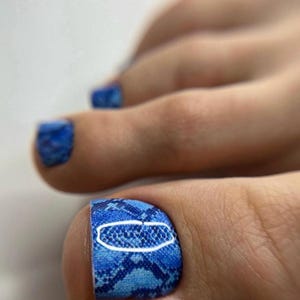 Può includere: Primo piano di un piede con unghie blu. L'alluce presenta un motivo a pelle di serpente blu con un disegno ovale bianco. Anche le altre unghie dei piedi sono dipinte di blu.