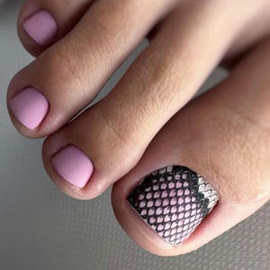 Peut inclure: Gros plan sur un pied avec une pédicure. Les ongles des orteils sont peints en rose clair mat, avec un orteil orné d'un motif peau de serpent noir, rose et blanc. Le teint est clair.