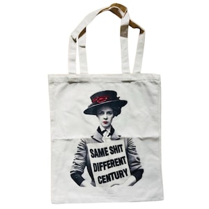 Könnte beinhalten: Weißer Stoffbeutel mit einem Aufdruck einer Frau mit Hut, die ein Schild mit der Aufschrift "SAME SHIT DIFFERENT CENTURY" hält. Die Tasche hat zwei Henkel und ist aus einem leinwandartigen Material.