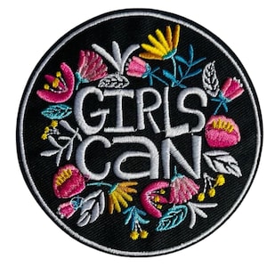 Könnte beinhalten: Runder, gestickter Aufnäher mit dem weißen Schriftzug "GIRLS CAN", umgeben von bunten floralen Designs. Der Aufnäher hat einen schwarzen Hintergrund und einen weißen Rand. Die Blumen sind rosa, gelb und blau.