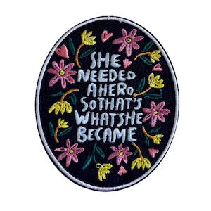 Könnte beinhalten: Ein ovaler, bestickter Aufnäher mit schwarzem Hintergrund und dem hellblauen Text SHE NEEDED A HERO SO THAT'S WHAT SHE BECAME. Das Design ist von bunten, gestickten Blumen in Rosa- und Gelbtönen sowie grünen Blättern umgeben.