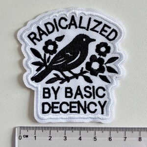 May include: A white embroidered patch with black thread detailing a bird perched on a branch surrounded by flowers. The text RADICALIZED BY BASIC DECENCY is stitched in black. A ruler at the bottom shows the patch is approximately 9.5 cm wide.