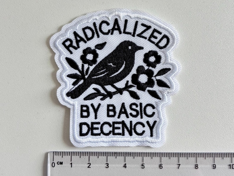 May include: A white embroidered patch with black thread detailing a bird perched on a branch surrounded by flowers. The text RADICALIZED BY BASIC DECENCY is stitched in black. A ruler at the bottom shows the patch is approximately 9.5 cm wide.