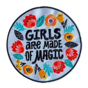 Könnte beinhalten: Runder, gestickter Aufnäher mit dem Text "GIRLS are MADE OF MAGIC" in schwarzer Schrift. Das Design ist von einem floralen Kranz mit rot, orange, gelb und türkis gestickten Blumen und Blättern umgeben.