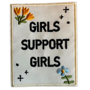 Könnte beinhalten: Gestickter Aufnäher mit dem Text "GIRLS SUPPORT GIRLS" in Schwarz auf weißem Hintergrund. Der Aufnäher zeigt gestickte Blumen und Pluszeichen. Der Rand ist hellbeige.