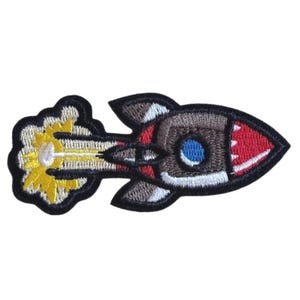 Könnte beinhalten: Ein gestickter Aufnäher in Form einer Rakete mit schwarzer Umrandung. Die Rakete hat eine rote Spitze mit weißen Zähnen, einen braunen Körper, ein blaues rundes Fenster und dunkelgraue Flossen. Eine gelb-weiße Abgaswolke zieht sich dahinter.