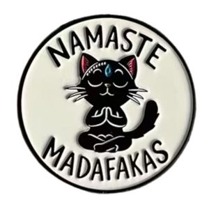 Könnte beinhalten: Runder Ansteck-Pin mit einer schwarzen Katze in Yoga-Pose, mit dem Text "NAMASTE MADAF*KAS" in schwarzer Schrift auf weißem Hintergrund. Die Katze hat ein drittes Auge und wird von einem schwarzen Rand umgeben.