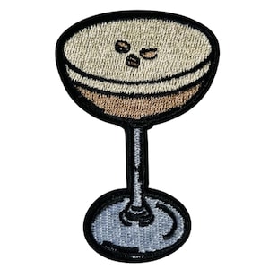 Könnte beinhalten: Gestickter Aufnäher mit einem Cocktail in einem Stielglas. Das Getränk hat eine hellbraune Oberseite mit drei dunkelbraunen Kaffeebohnen, eine braune Mittelschicht und eine schwarze Umrandung. Stiel und Fuß sind schwarz und grau.