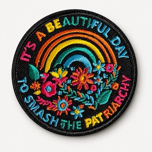 Könnte beinhalten: Ein runder, schwarzer Aufnäher mit einer bunten Regenbogenstickerei und verschiedenen Blumen in Rosa, Blau, Gelb und Grün. Der Text "IT'S A BEAUTIFUL DAY TO SMASH THE PATRIARCHY" ist in leuchtenden Farben um das Motiv herum eingestickt.