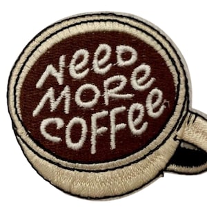 Könnte beinhalten: Gestickter Aufnäher in Form einer Kaffeetasse mit den Worten "Need More Coffee" in weißer Schrift auf braunem Hintergrund. Die Tasse hat einen beigen Rand und Henkel mit schwarzer Umrandung.