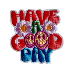 Könnte beinhalten: Ein gestickter Aufnäher mit dem Text HAVE A GOOD DAY in kräftigen roten und blauen Buchstaben. Das Design enthält zwei lila Blumen mit gelben Mitten und zwei gelbe Smileys mit rosa Umrandungen in einer farbenfrohen Anordnung.