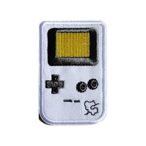 Könnte beinhalten: Ein rechteckiger Stickaufnäher mit dem Design einer weißen tragbaren Spielkonsole. Das Gerät verfügt über einen gelben Bildschirm mit schwarzem Rand, ein schwarzes Steuerkreuz, zwei schwarze runde Tasten und kleine schwarze Liniendetails.